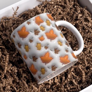May include: White ceramic mug with a repeating pattern of orange, yellow, and brown fall leaves. The mug has a white handle and is nestled in brown packing material inside a white box. The mug is a fall-themed item.