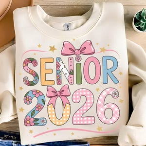 May include: Cream-colored sweatshirt with the words "SENIOR 2026" in colorful, floral, and gingham patterns. The design includes pink bows and gold star accents, perfect for a graduation celebration.