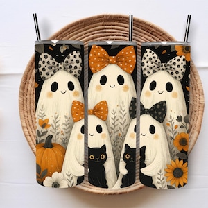 May include: Three Halloween-themed tumblers with black straws, featuring smiling ghost characters with bowties and black cats. The design includes pumpkins, sunflowers, and floral accents on a black background. The tumblers are set on a woven tray.