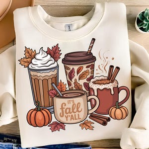 Könnte beinhalten: Cremefarbenes Sweatshirt mit einem Herbst-Themen-Design. Die Grafik zeigt Kaffeegetränke, Kürbisse, Herbstblätter und Zimtstangen. Der Satz "It's Fall Y'all" ist auf einer Tasse aufgedruckt. Ein gemütliches Kleidungsstück zum Thema Herbst.