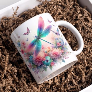May include: White ceramic mug featuring a watercolor design of a dragonfly with pink, blue, and green wings, surrounded by flowers and butterflies. The mug has a white handle and is presented in a gift box with brown paper filler.