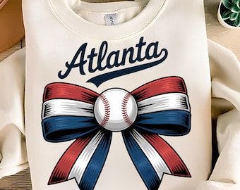 May include: Cream-colored sweatshirt featuring the word "Atlanta" in navy blue script above a baseball-themed design. The design includes a baseball in the center of a red, white, and blue ribbon bow.