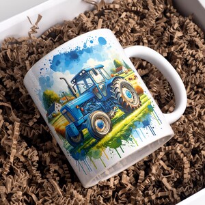 Op de afbeelding: Witte keramische mok met een aquarel schilderij van een blauwe tractor in een veld. De mok heeft een wit handvat en wordt gepresenteerd in een witte doos met bruin papier. Het tractorontwerp bevat een blauwe lucht en groen gras.