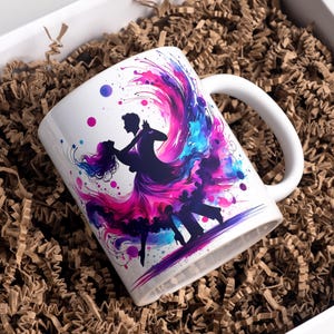 May include: White ceramic mug featuring a silhouette of a dancing couple with a vibrant watercolor design. The design incorporates shades of pink, purple, and blue, creating a dynamic and artistic look. The mug is placed in a white box with brown packing material.