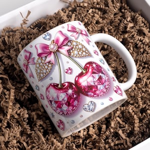 Puede incluir: Taza de cerámica blanca con un diseño de dos cerezas con facetas rosas en forma de joya y detalles dorados. Las cerezas están adornadas con lazos rosas y adornos con forma de diamantes. La taza está en una caja blanca con relleno de papel marrón.