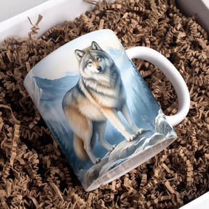 May include: White ceramic mug featuring a detailed illustration of a wolf standing on a rock with a mountain backdrop. The mug has a white handle and is presented in a gift box with brown paper filler.