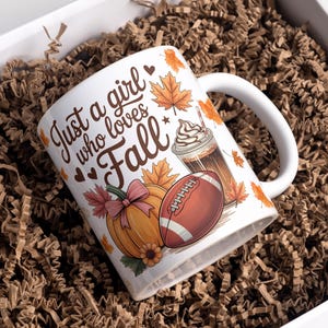 Könnte beinhalten: Weiße Keramiktasse mit dem Text "Just a girl who loves Fall", einem Kürbis, einem Football und einem Kaffeegetränk. Die Tasse befindet sich in einer weißen Box, die mit braunem Verpackungsmaterial gefüllt ist.
