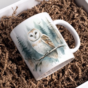 May include: White ceramic mug featuring a watercolor illustration of a barn owl perched on a branch in a forest setting. The mug has a white handle and is presented in a gift box with brown crinkle paper.