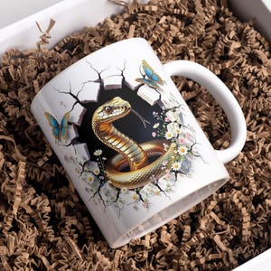 Może przedstawiać: Biały ceramiczny kubek z szczegółową ilustracją węża wychodzącego z pękniętej ściany, otoczonego motylami i kwiatami. Kubek znajduje się w białym pudełku prezentowym wypełnionym brązowym materiałem opakowaniowym.