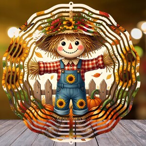 Op de afbeelding: Een ronde metalen windspinner met een kleurrijke illustratie van een vogelverschrikker in een rood geruit overhemd en een blauwe tuinbroek met zonnebloemaccenten. Het ontwerp bevat zonnebloemen, oranje pompoenen en vlinders bij een hek.