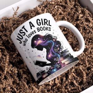Puede incluir: Taza de cerámica blanca con el texto "JUST A GIRL who loves BOOKS". La taza presenta la silueta de una persona leyendo, con un diseño de galaxia en su cabello y libros. La taza está en una caja blanca con material de embalaje marrón.