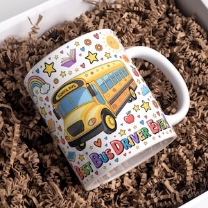 Op de afbeelding: Witte keramische mok met een kleurrijk schoolbusontwerp, de tekst "BEST BUS DRIVER EVER!" en andere illustraties. De mok zit in een witte doos met bruin verpakkingsmateriaal.