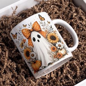 May include: A white ceramic mug featuring a printed illustration of a ghost wearing a large orange polka-dot bow, surrounded by sunflowers, daisies, pumpkins, and monarch butterflies. The mug has a curved handle and a smooth, glossy finish.