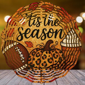 Op de afbeelding: Ronde herfstdecoratie met de tekst "'tis the season". Het heeft een voetbal, een pompoen met een luipaardprint en een pumpkin spice latte. De achtergrond heeft een wazig bokeh-effect.