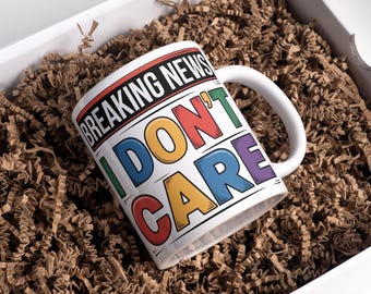 Könnte beinhalten: Weiße Keramiktasse mit dem Text "BREAKING NEWS I DON'T CARE" in einer farbenfrohen, fetten Schriftart. Die Tasse befindet sich in einer weißen Box, die mit braunem Papier gefüllt ist.