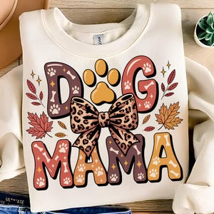 May include: Cream-coloured sweatshirt featuring the text "DOG MAMA" in a decorative font. The design incorporates paw prints, bones, a leopard print bow, and autumn leaves. A comfortable and fashionable top for dog enthusiasts.