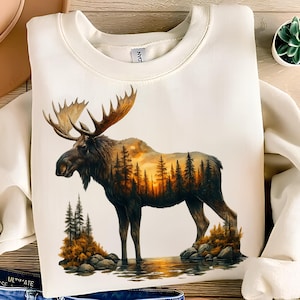 May include: Cream-colored sweatshirt featuring a detailed illustration of a moose with large antlers. The moose's body incorporates a sunset scene with trees. The design is set against a water reflection, creating a nature-themed aesthetic.