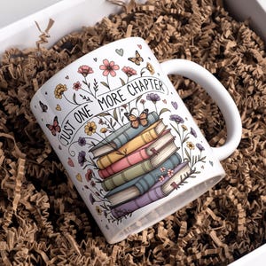 May include: White ceramic mug featuring a stack of colourful books, flowers, and butterflies. The mug has the text "JUST ONE MORE CHAPTER" and is in a white box filled with brown packing material.