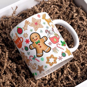 Puede incluir: Taza de cerámica blanca con un diseño navideño festivo. La taza presenta hombres de jengibre, bastones de caramelo, estrellas y otras ilustraciones temáticas navideñas. La taza está colocada dentro de una caja blanca llena de virutas de papel marrón.