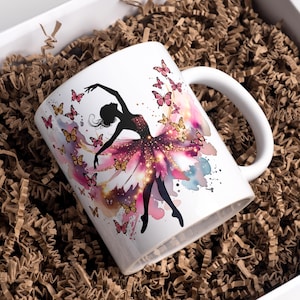Puede incluir: Taza de cerámica blanca con una silueta de bailarina en un vestido de acuarela rosa y morado, rodeada de mariposas. La taza tiene un asa blanca y se presenta en una caja de regalo con relleno de papel marrón.