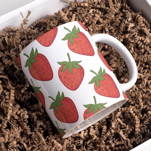 Puede incluir: Taza de cerámica blanca con un patrón repetido de fresas rojas con hojas verdes. La taza está dentro de una caja blanca llena de relleno de papel marrón.