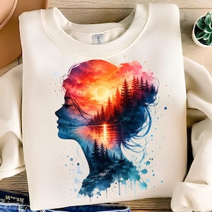 Könnte beinhalten: Ein cremefarbenes Sweatshirt mit Rundhalsausschnitt und einem Grafikdruck, der das Seitenprofil eines Kopfes zeigt. Die Silhouette ist mit einer lebendigen Aquarell-Landschaft gefüllt, die einen Sonnenuntergang über einem See und Tannen zeigt.