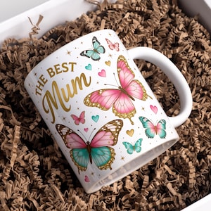 May include: White ceramic mug with the text "The Best Mum" in gold script, decorated with pink, teal, and brown butterflies and hearts. The mug is inside a white gift box filled with brown crinkle paper.