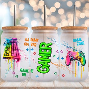 May include: Three glass tumblers with bamboo lids and metal straws. Each tumbler features a gaming theme with colorful graphics: a keyboard, the word "GAMER", and a game controller. The words "GAME ON" and "GAME ZONE" are also visible.