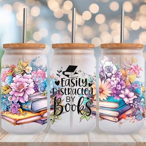 Puede incluir: Tres vasos de vidrio con tapas de madera y pajitas de metal. Cada vaso presenta un diseño floral con flores rosas, moradas y azules. El vaso central tiene el texto "Easily Distracted By Books".