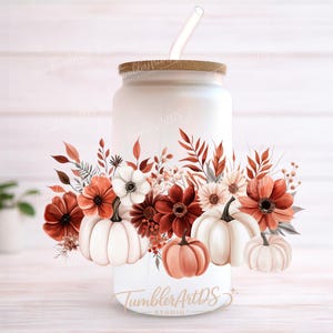 Puede incluir: Un vaso de vidrio blanco con tapa de madera y pajita transparente. El vaso está decorado con un arreglo floral de temática otoñal con calabazas, flores y hojas en tonos naranja, blanco y marrón. El texto "TumblerArtDS Studio" es visible.