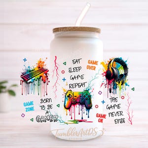 Puede incluir: Vaso de vidrio blanco con tapa de madera y pajita de vidrio. El vaso presenta gráficos coloridos de un teclado, auriculares y mandos de juego con el texto "Eat Sleep Game Repeat" y "Born to be a Gamer".