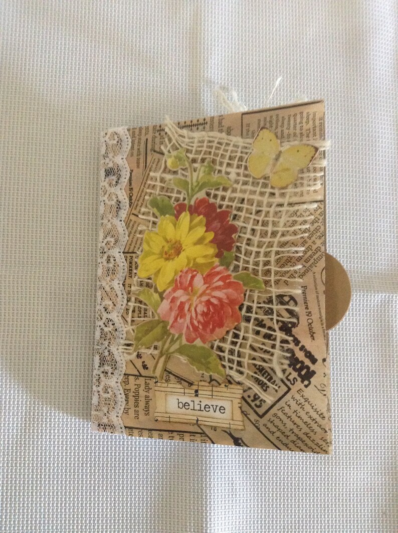 Puede incluir: Una tarjeta hecha a mano con un fondo de arpillera y encaje. La tarjeta presenta un dise&ntilde;o floral con una flor amarilla, roja y rosa. La tarjeta tambi&eacute;n tiene una mariposa y la palabra "creer" escrita en ella.