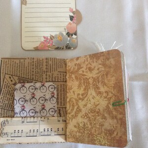Puede incluir: Un peque&ntilde;o diario de tela marr&oacute;n con una lista de tareas con estampado floral pegada a la portada. El diario est&aacute; abierto para revelar una p&aacute;gina con un fondo de papel estampado y un peque&ntilde;o trozo de partitura musical.