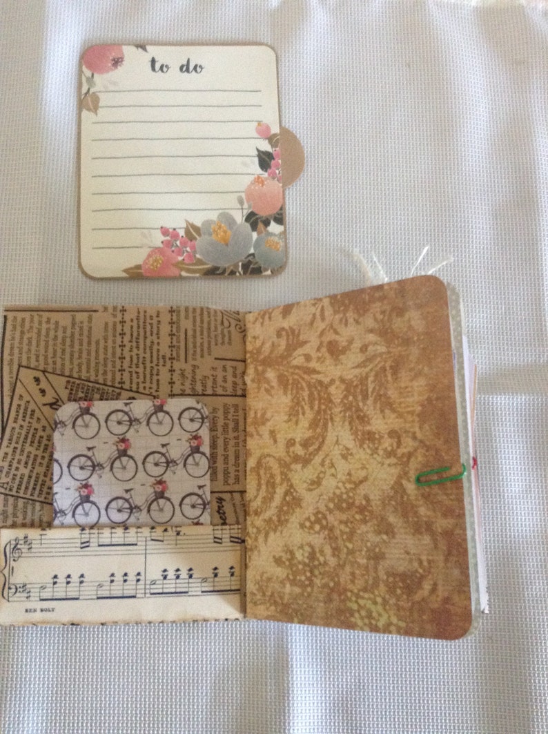 Puede incluir: Un peque&ntilde;o diario de tela marr&oacute;n con una lista de tareas con estampado floral pegada a la portada. El diario est&aacute; abierto para revelar una p&aacute;gina con un fondo de papel estampado y un peque&ntilde;o trozo de partitura musical.