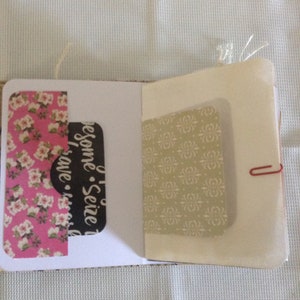 Puede incluir: Un peque&ntilde;o cuaderno blanco con un papel estampado floral rosa y blanco en el lado izquierdo, un papel negro con el texto "Seize the Awesome - Have a Nice Day" en el lado derecho y un papel estampado beige en la parte posterior. El cuaderno est&aacute; cerrado y tiene un clip de papel rojo que lo mantiene unido.
