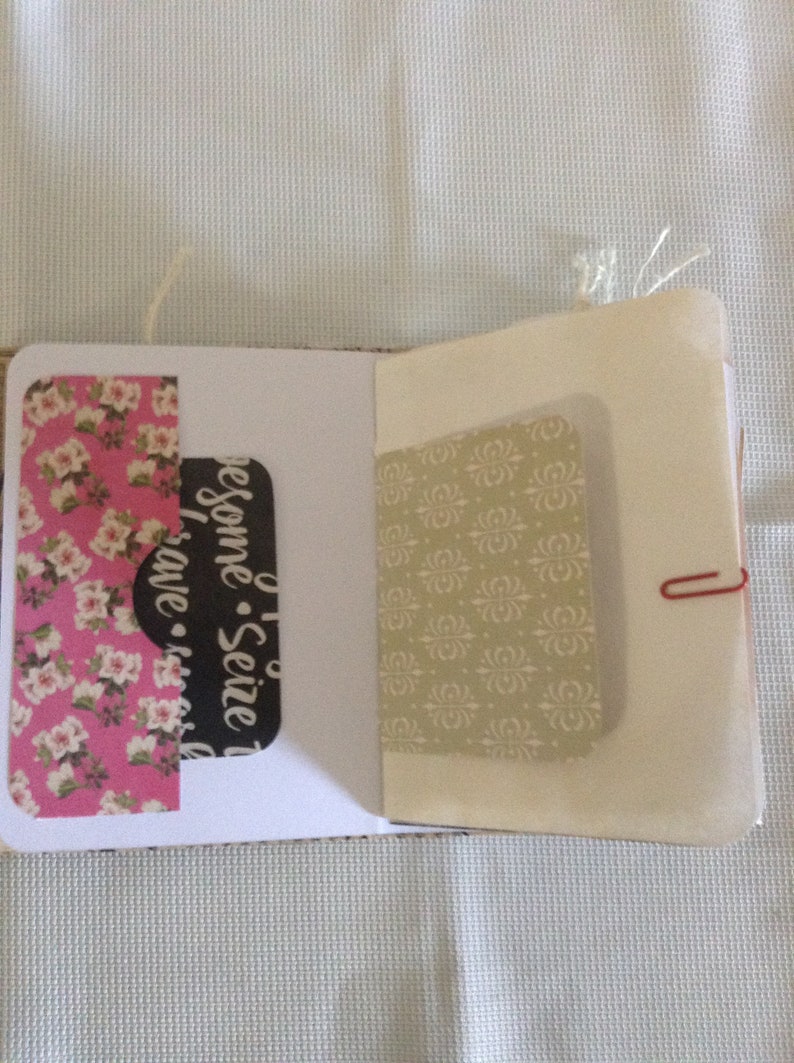 Puede incluir: Un peque&ntilde;o cuaderno blanco con un papel estampado floral rosa y blanco en el lado izquierdo, un papel negro con el texto "Seize the Awesome - Have a Nice Day" en el lado derecho y un papel estampado beige en la parte posterior. El cuaderno est&aacute; cerrado y tiene un clip de papel rojo que lo mantiene unido.
