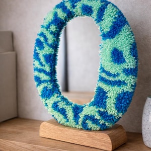 Peut inclure: Miroir ovale avec un cadre texturé et touffeté dans les tons de bleu et de vert. Le miroir repose sur une petite base en bois naturel, ajoutant une touche de style organique. Design moderne et décoratif.