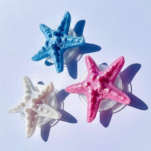 May include: Three starfish-shaped decorations in blue, pink, and white. Each starfish is mounted on a clear, circular base. They are arranged on a white surface, casting shadows. The starfish have a textured surface. Ideal for coastal decor.