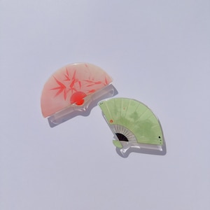 May include: Two decorative fan-shaped objects. One is light pink with red bamboo and a red circle design. The other is light green with a black and white fan design and a small orange circle. Both have a clear handle.