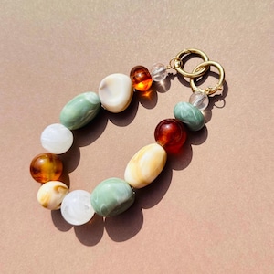 May include: A colourful beaded bracelet with green, white, amber, and cream beads. The bracelet is connected with gold-coloured metal rings and clasps. The beads are oval and round, creating a unique accessory. The bracelet is approximately 18 cm in length.