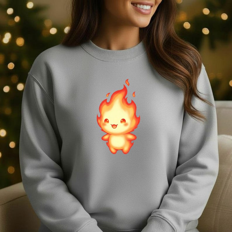 May include: A grey sweatshirt with a cartoon fire character printed on the front. The fire character is orange and yellow with a smiling face. The sweatshirt has a crew neck and is a solid colour.