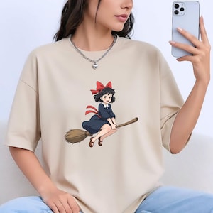 May include: Beige t-shirt with a cartoon illustration of a young witch on a broom. The witch has black hair, a red bow, and a dark blue dress. The broom is brown, and the design has a whimsical style.