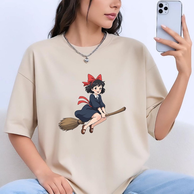 May include: Beige t-shirt with a cartoon illustration of a young witch on a broom. The witch has black hair, a red bow, and a dark blue dress. The broom is brown, and the design has a whimsical style.