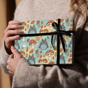 Puede incluir: Una caja de regalo envuelta en papel con un estampado de personajes de dibujos animados, setas y hojas de oto&ntilde;o en tonos azules, naranjas y amarillos. Est&aacute; atada con una cinta de terciopelo negro.