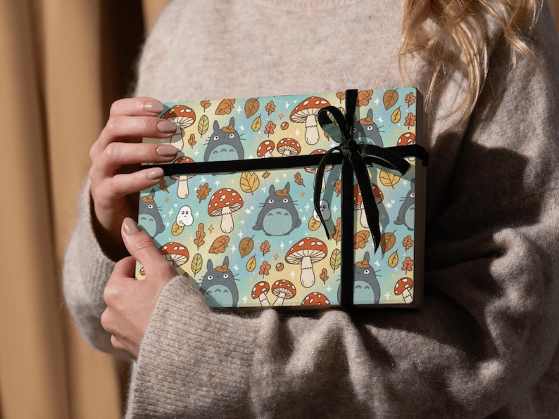 Puede incluir: Una caja de regalo envuelta en papel con un estampado de personajes de dibujos animados, setas y hojas de oto&ntilde;o en tonos azules, naranjas y amarillos. Est&aacute; atada con una cinta de terciopelo negro.