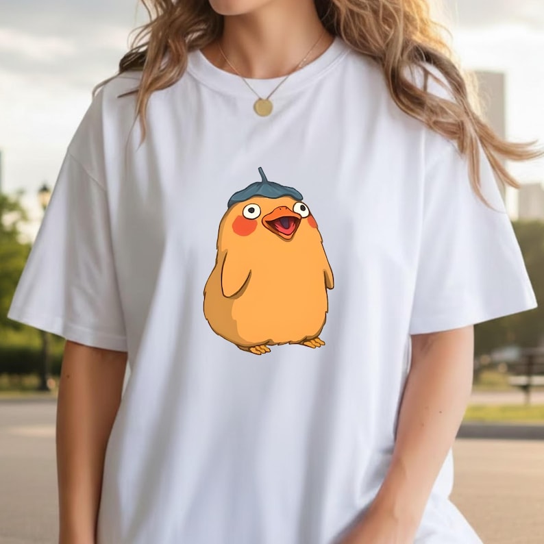 May include: White t-shirt with a cartoon illustration. The design features a yellow, round creature with a blue hat, red cheeks, and an open beak. The graphic is centrally placed, creating a fun and lighthearted look.