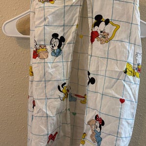 Puede incluir: Una prenda de tela blanca con un patrón de cuadrícula azul e ilustraciones coloridas de personajes de Disney, incluyendo a Mickey Mouse, Minnie Mouse y Pluto. El artículo tiene un pequeño lazo blanco en el centro superior y está en una percha.