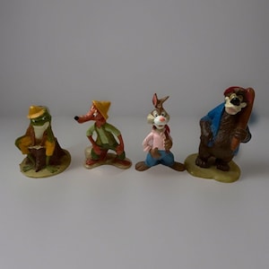 May include: Four small figurines of cartoon animals. A frog in a yellow hat and coat, a fox in a yellow hat and green shirt, a rabbit in a pink shirt and blue trousers, and a bear in a blue coat and red hat.