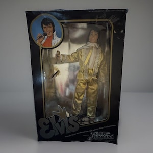 May include: An Elvis Presley doll in a gold jumpsuit with a white scarf, packaged in a black box. The box features an image of Elvis singing and the text "Elvis" and "Graceland". The doll includes a microphone and guitar.
