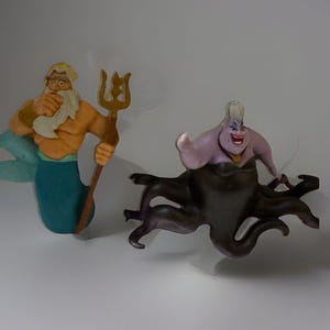 Puede incluir: Dos figuras de personajes de una película de fantasía. Uno es un tritón con cola turquesa, barba blanca y tridente dorado. El otro es una bruja marina de piel púrpura con tentáculos negros.